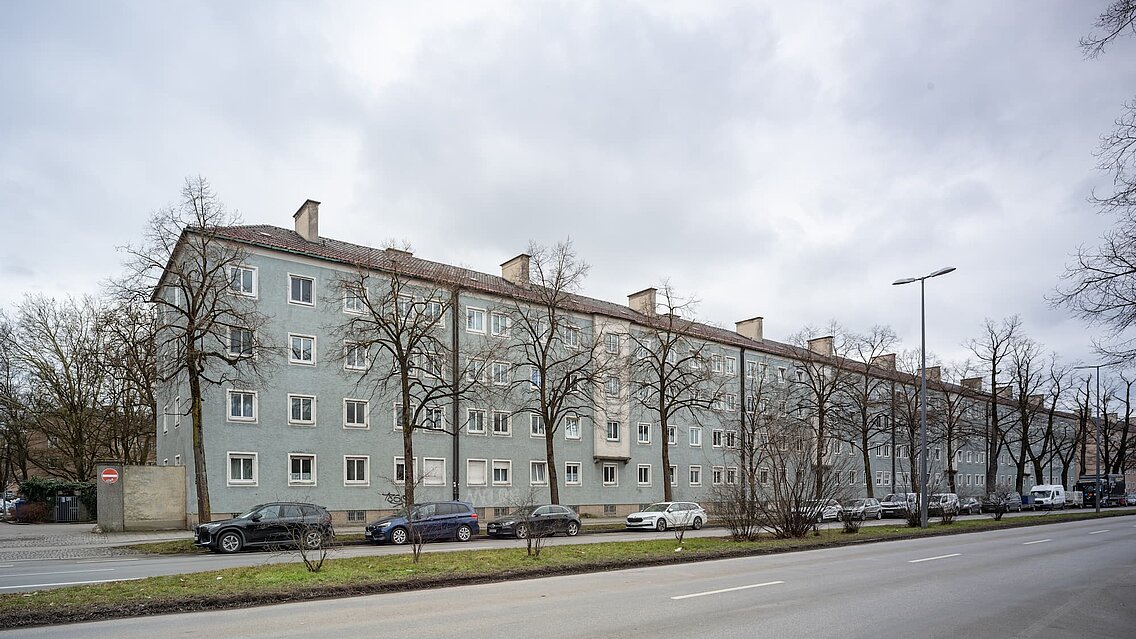 Mehrgeschossiges Wohngebäude mit grauer Fassade, mehreren Fenstern und parkenden Autos vor einer Straße.